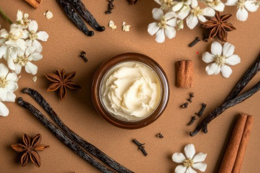 WARMFIELDS Lavender Vanilla Body Butter in a brown jar surrounded by vanilla beans, cinnamon sticks, star anise, and white flowers. Ideal for deep skin moisture.